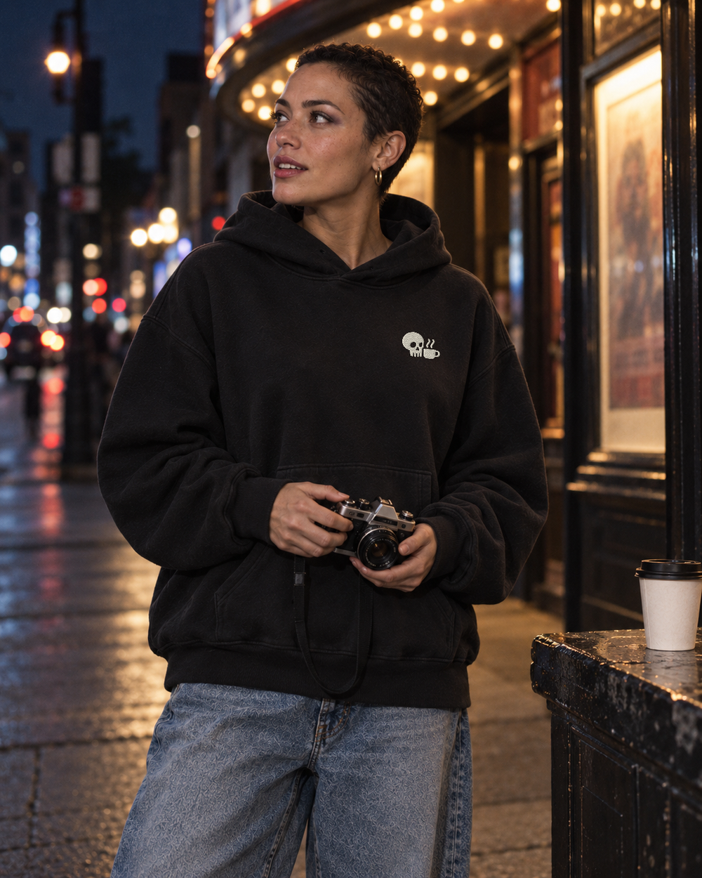 A woman holds a camera outside a lit cinema wearing the Vintage Black Coffee Companion Hoodie, with a small white skull-and-cup mark on the left chest, and faded blue jeans.
