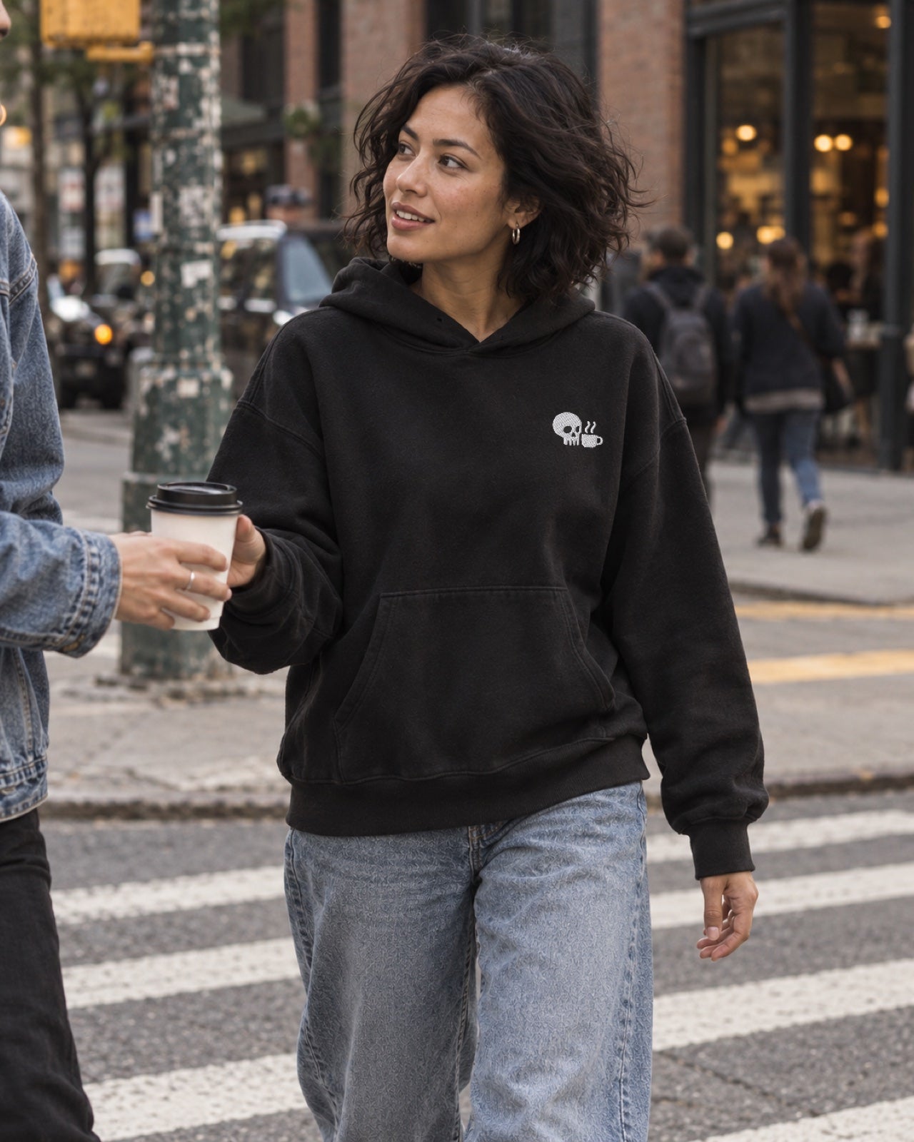 Editorial study of a woman in an SDC black hoodie accepting a takeaway coffee on a city street